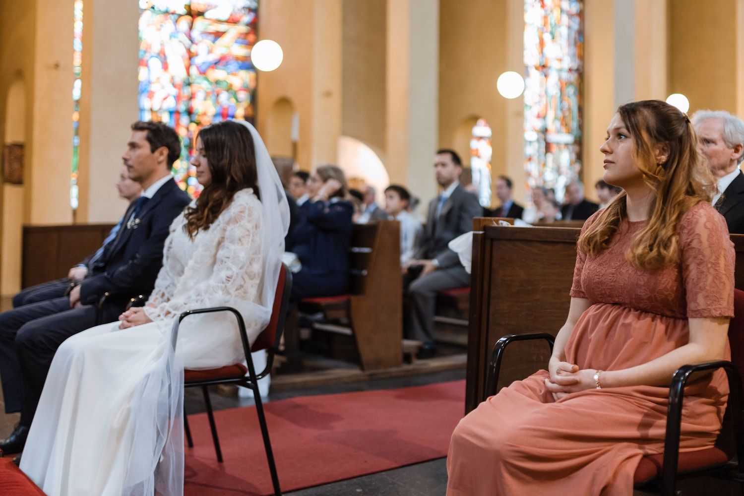 Wedding guests seated in church pews react emotionally during ceremony with stained glass windows visible in background.
