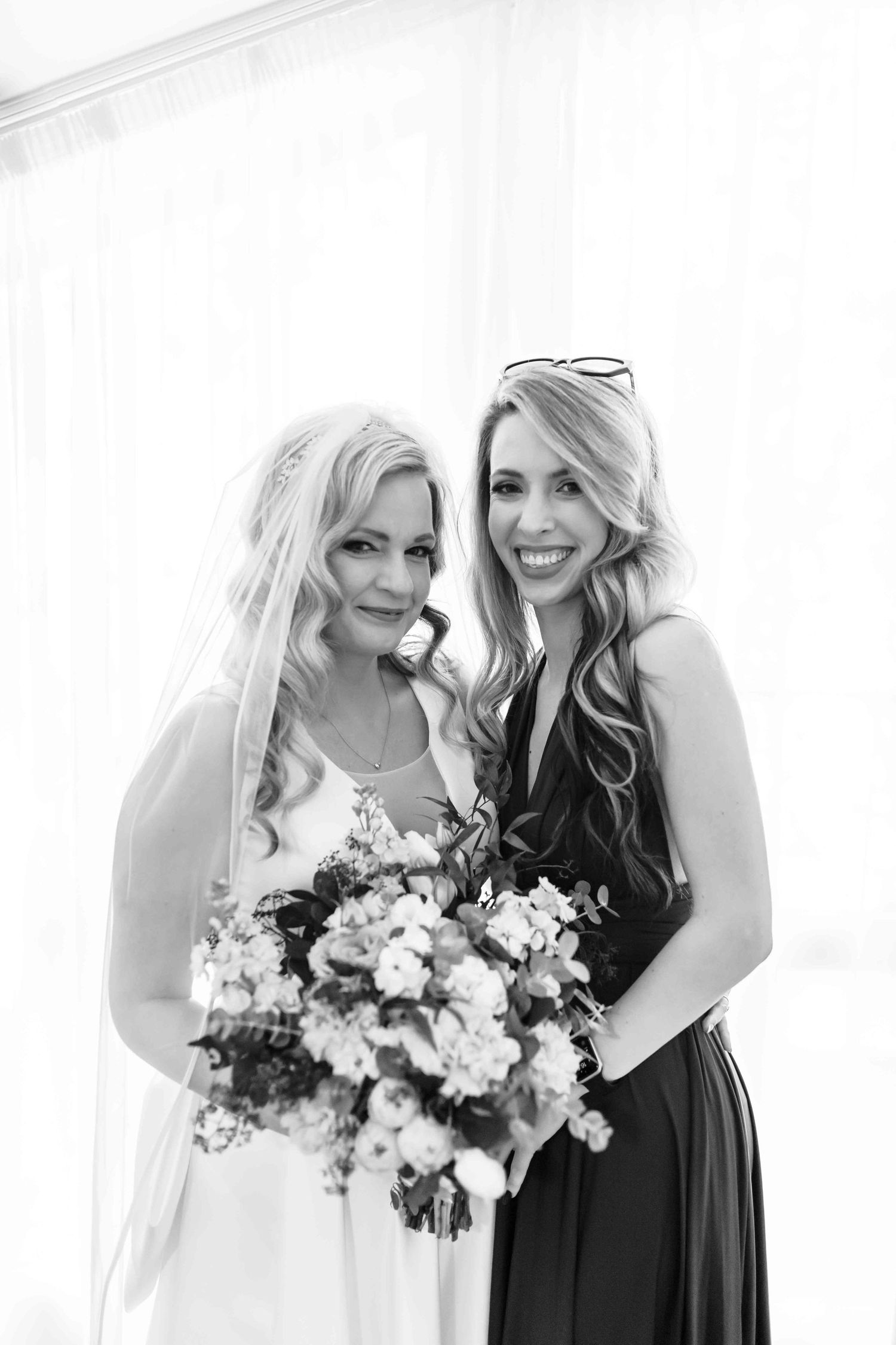 Two women in black and white dresses share joyful moments while holding floral bouquets during a wedding photoshoot.