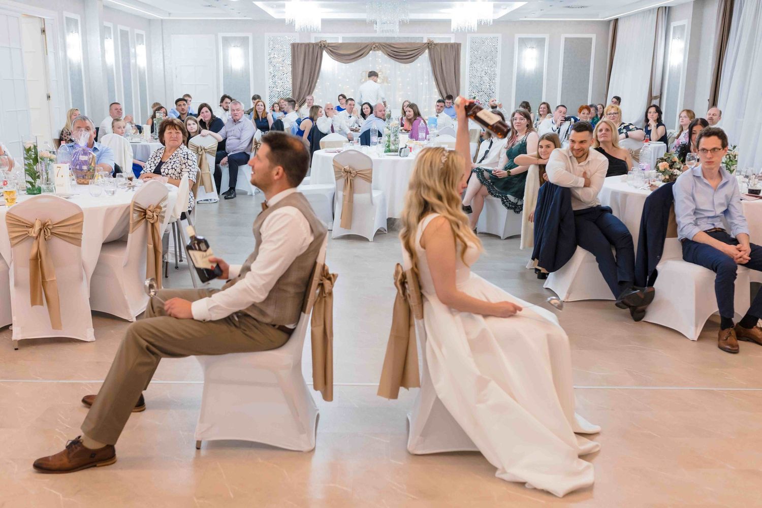Wedding guests laugh and cheer during a fun reception game where a couple sits back-to-back in chairs.