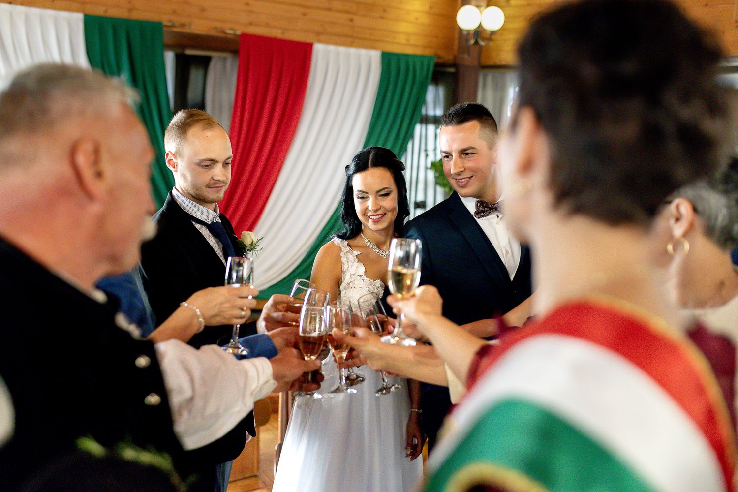 Wedding guests raise glasses in a toast during a celebration with Italian flag-colored decorations in the background.