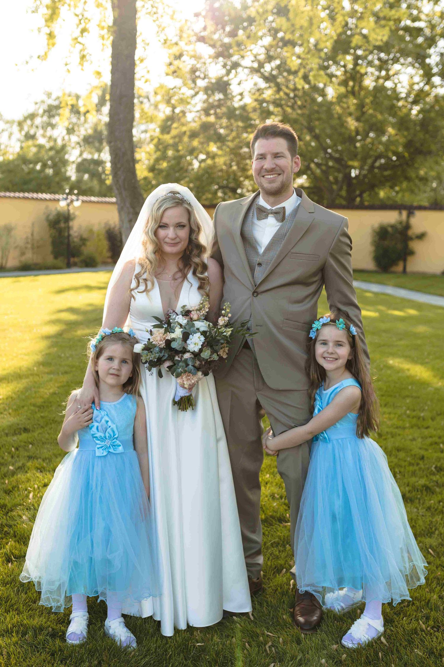 Wedding party in white dress and gray suit poses with two flower girls wearing bright blue tulle dresses in garden setting.