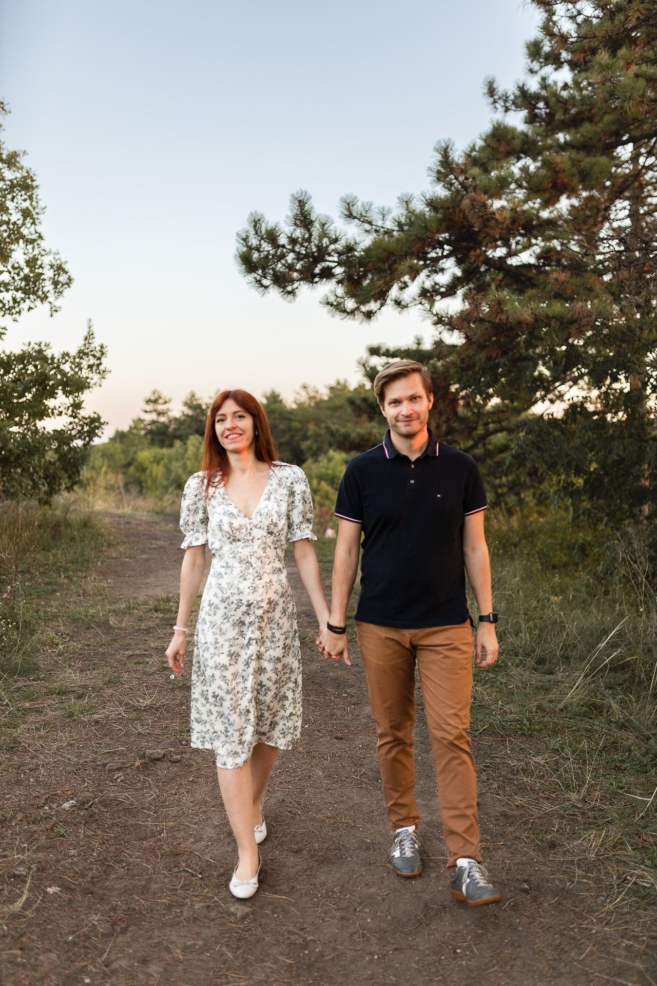 Couple walking hand in hand along a dirt path with pine trees in the background.