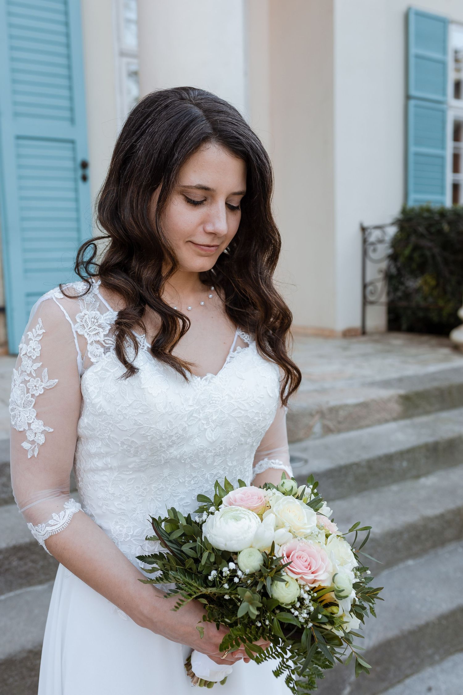 A bride in a white lace dress with sheer sleeves holds a romantic bouquet of white and pink flowers.