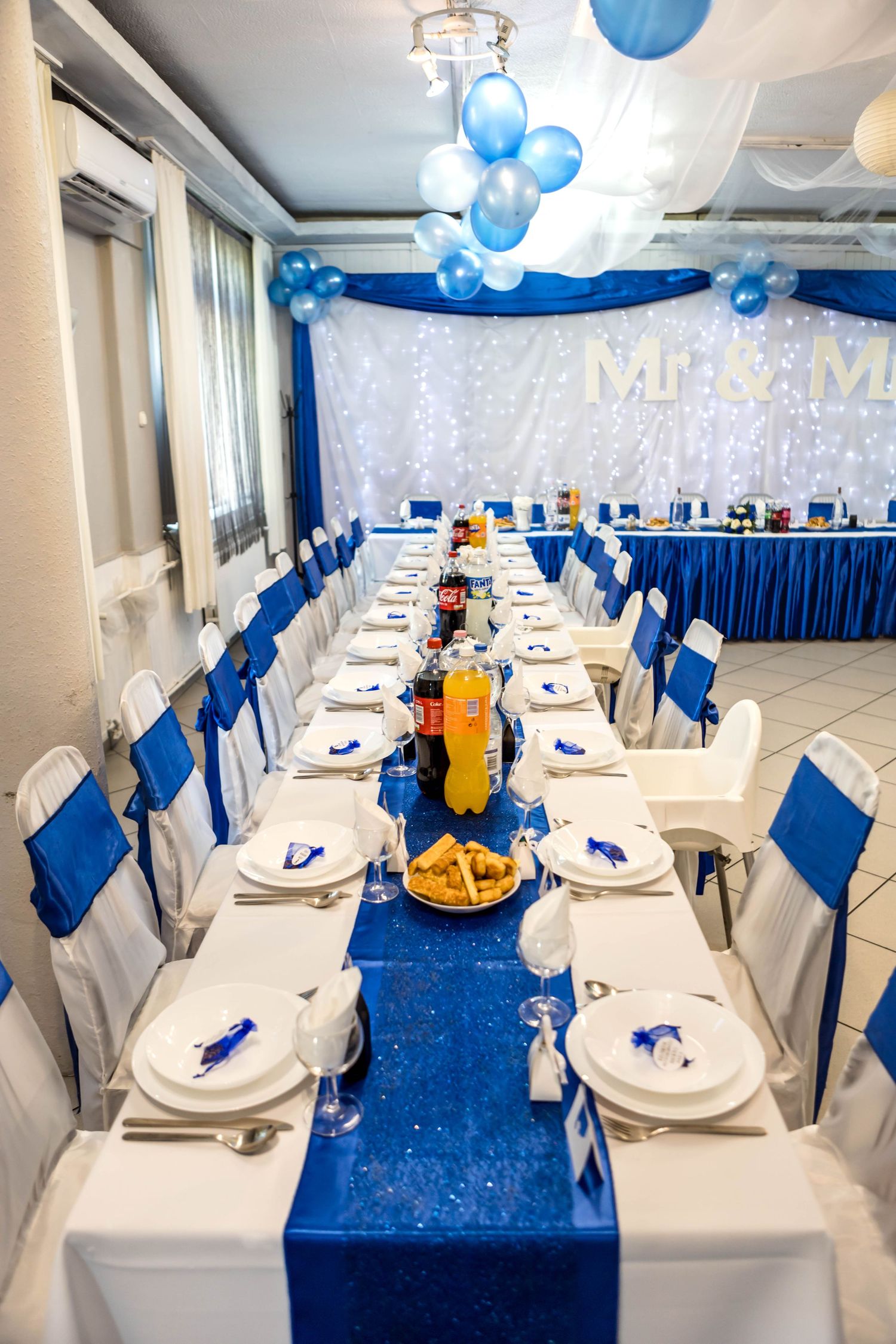 Elegant banquet hall table settings with royal blue and white decor featuring balloons and twinkle light backdrop.