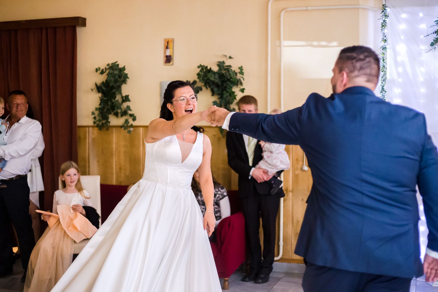 A couple shares their first dance at a wedding reception in a hall decorated with greenery and curtains.