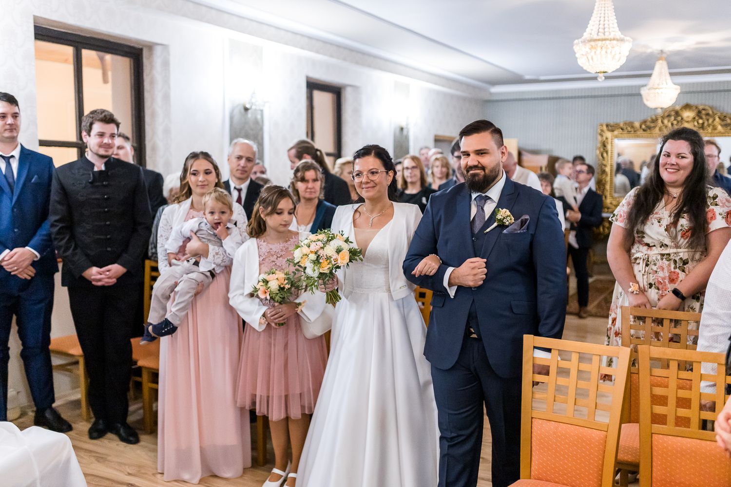 A wedding party stands in a church with bridesmaids in pink dresses and groomsmen in navy suits.