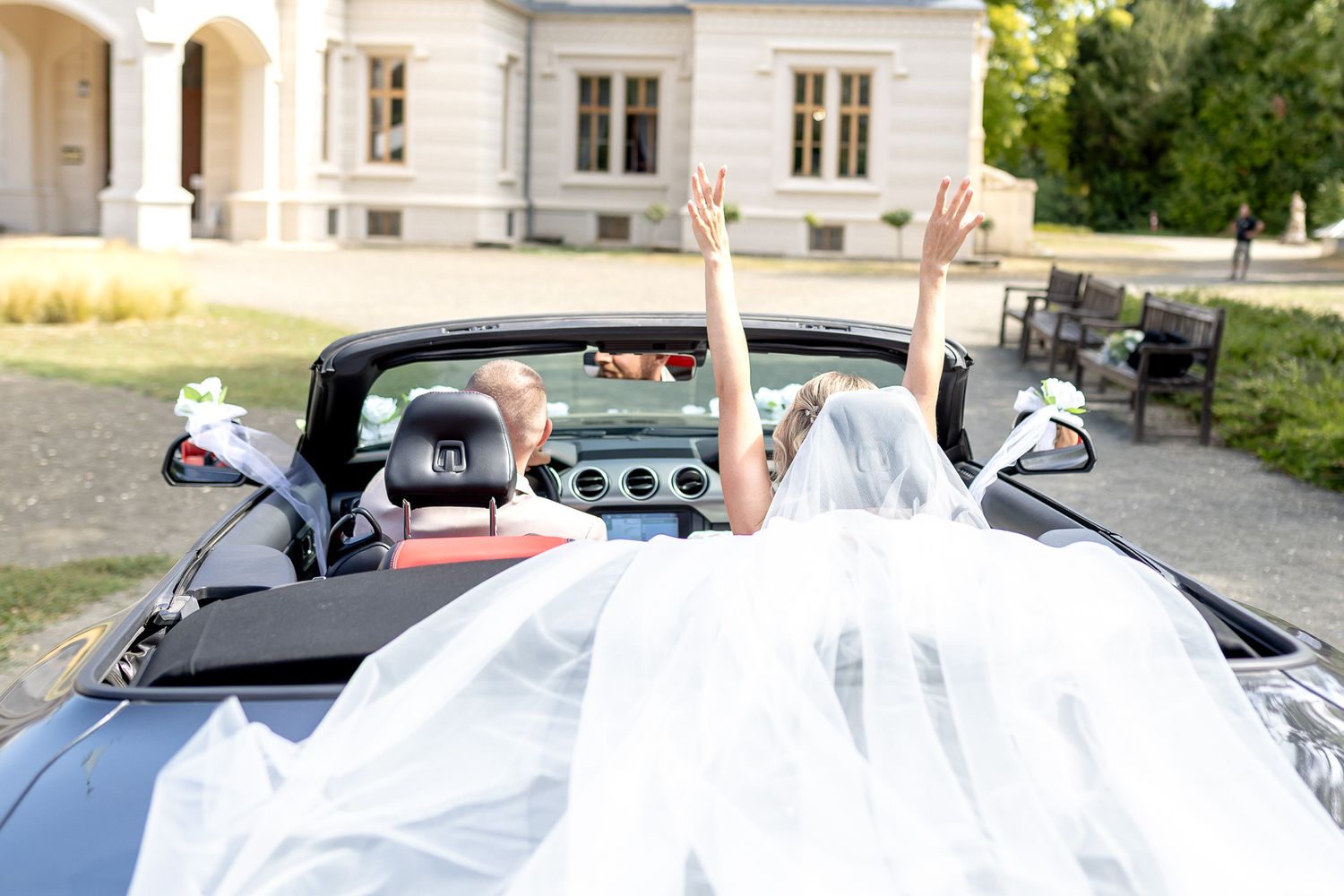 A wedding dress train flows elegantly over the back of a classic convertible car during a sunny outdoor celebration.