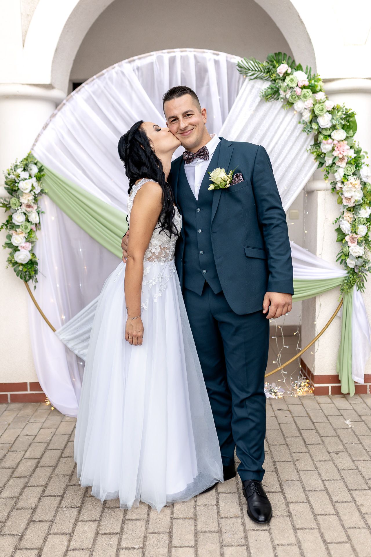Wedding couple poses together on stone patio with white and green floral arch decorations in the background.