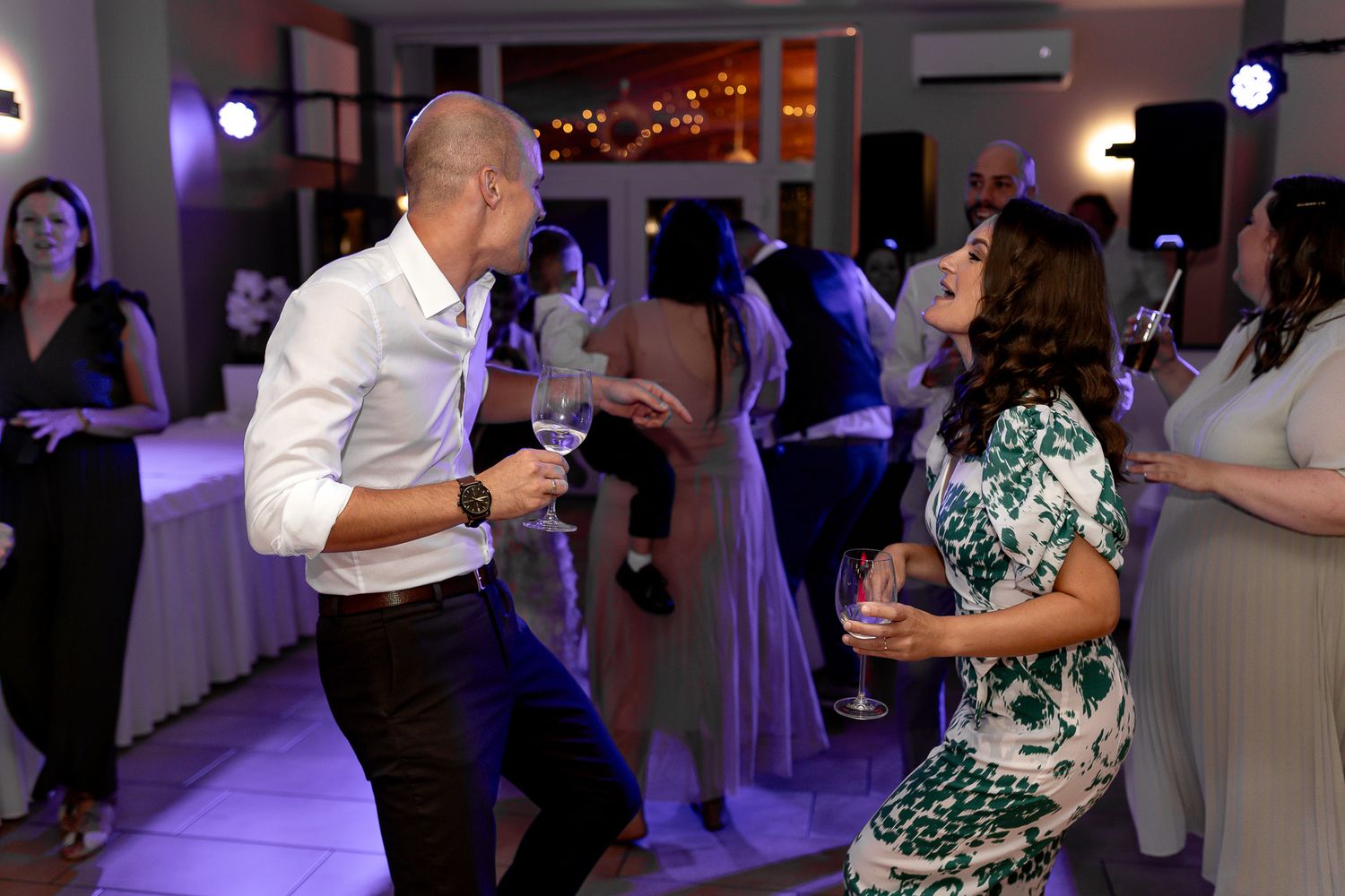 Couples dancing together at an indoor wedding reception with purple mood lighting and white decor.
