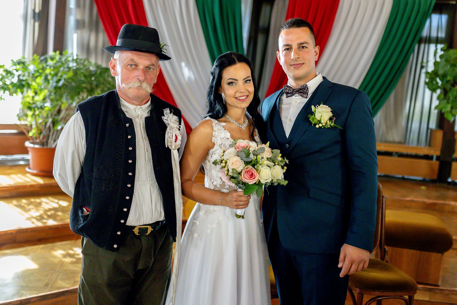 A wedding party poses in front of red and green draped curtains at an elegant indoor venue.