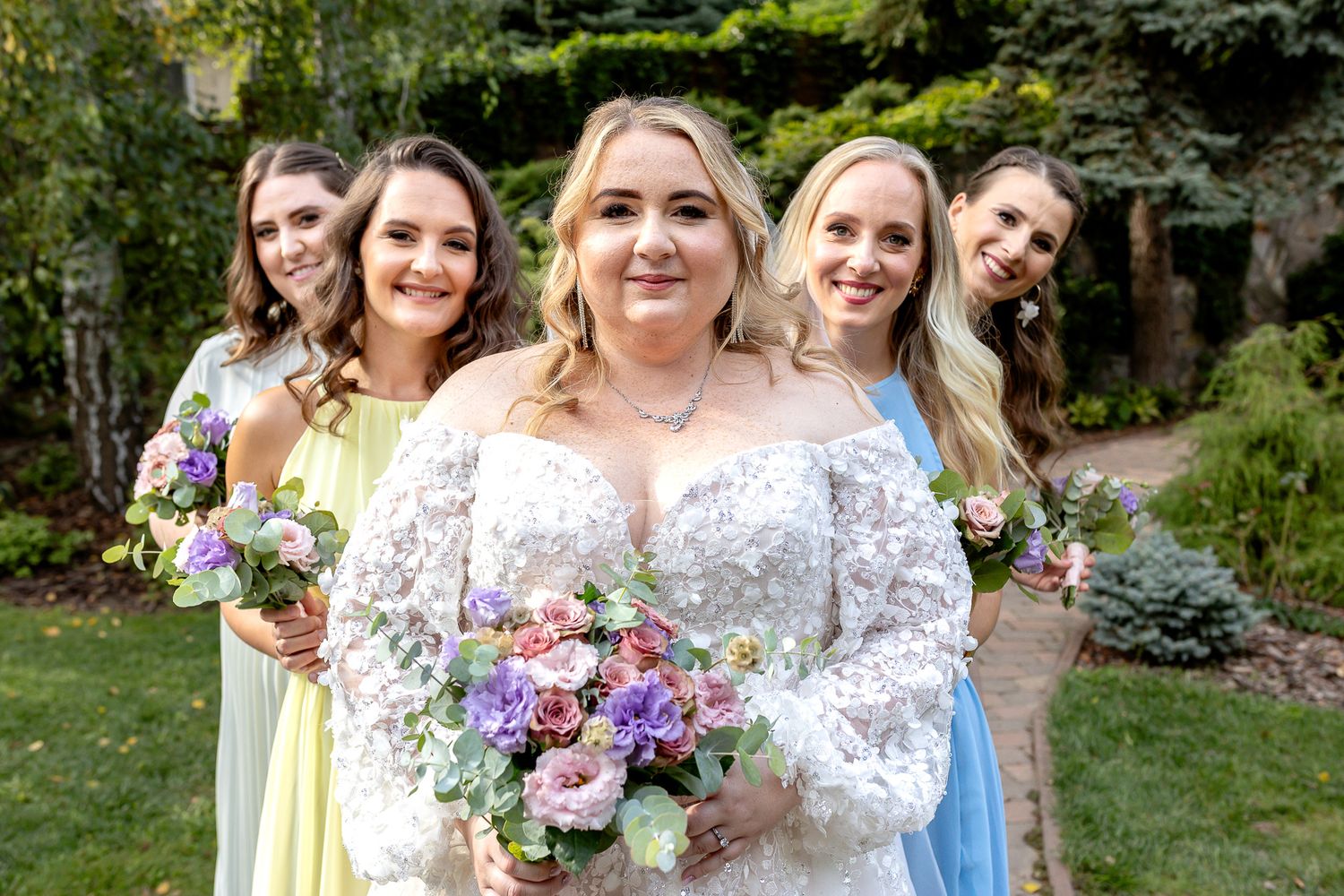 Wedding party poses together in garden wearing pastel dresses and holding floral bouquets.