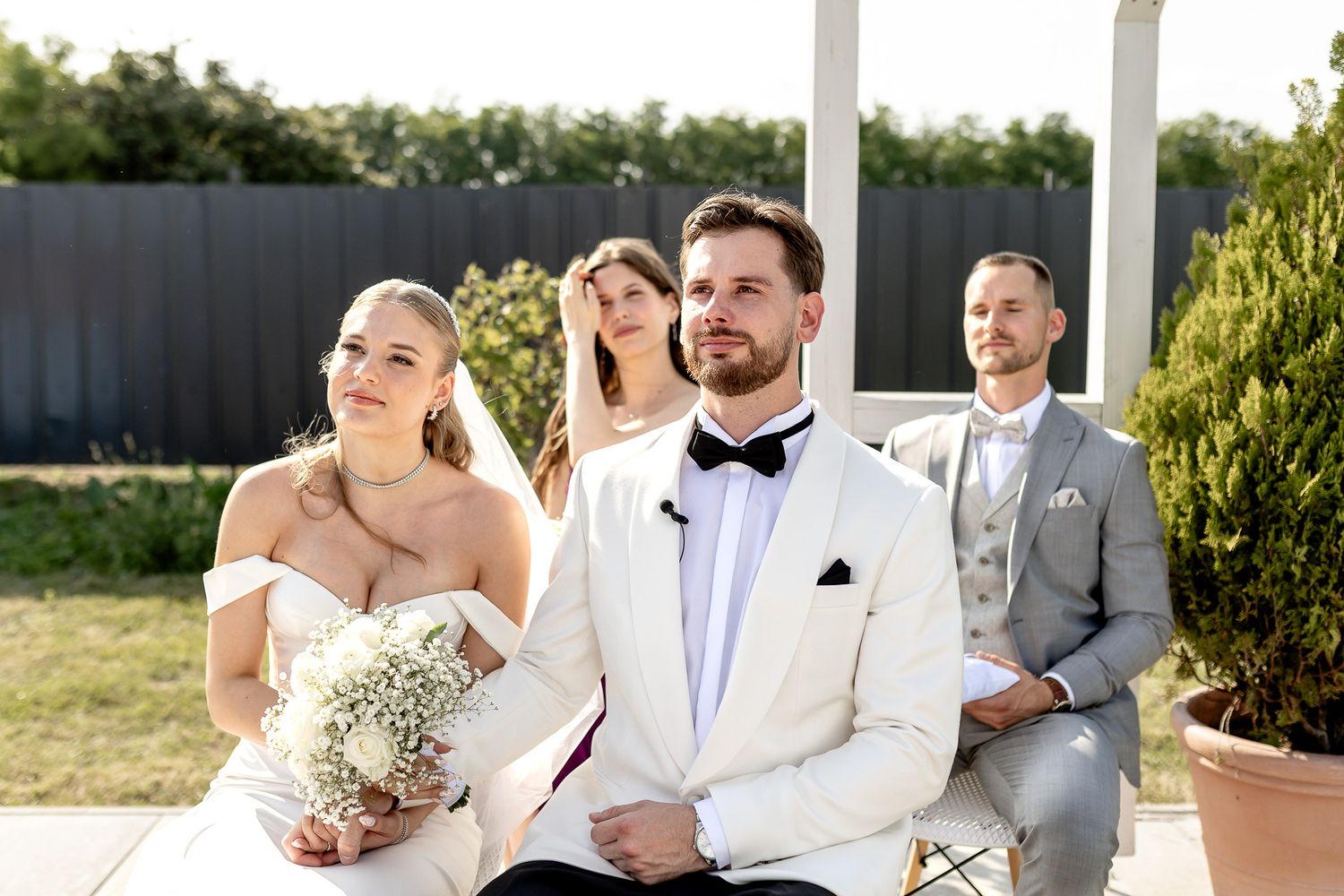 Wedding ceremony participants stand in line under white outdoor pergola with greenery.