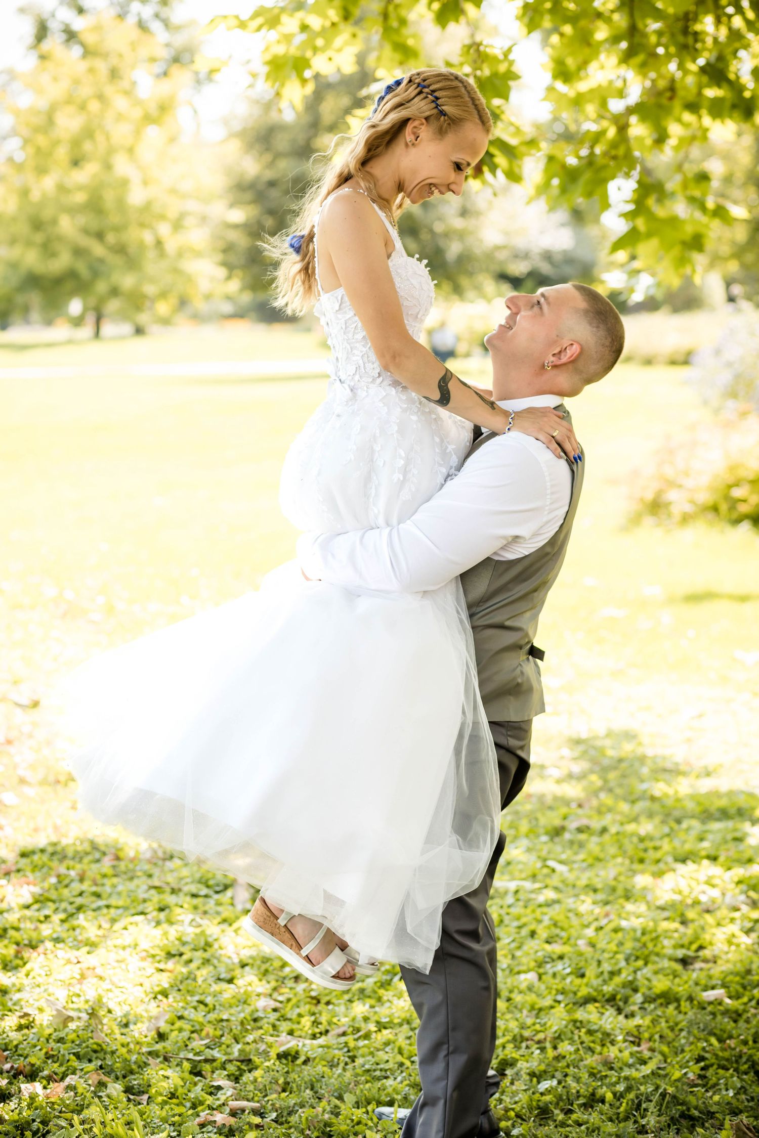 A romantic wedding couple embraces in a sun-dappled garden setting with lush greenery in the background.