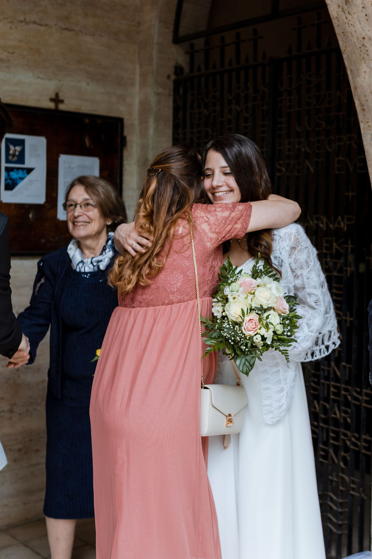 A bride in white dress and bridesmaid in coral pink dress share joyful moments together in a stone hallway.
