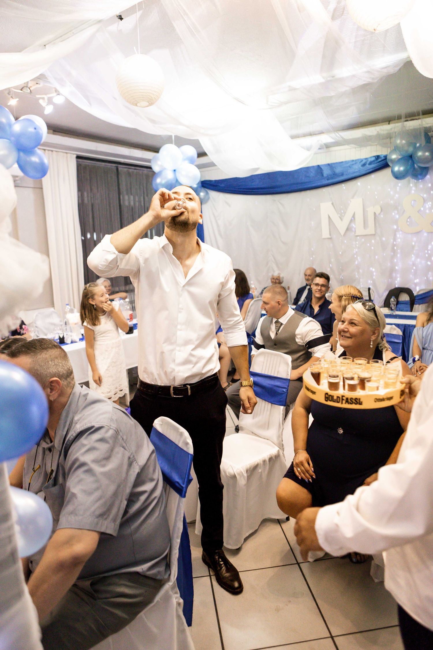 Wedding guests dance and celebrate with balloons and decorations in a white and blue themed reception hall.