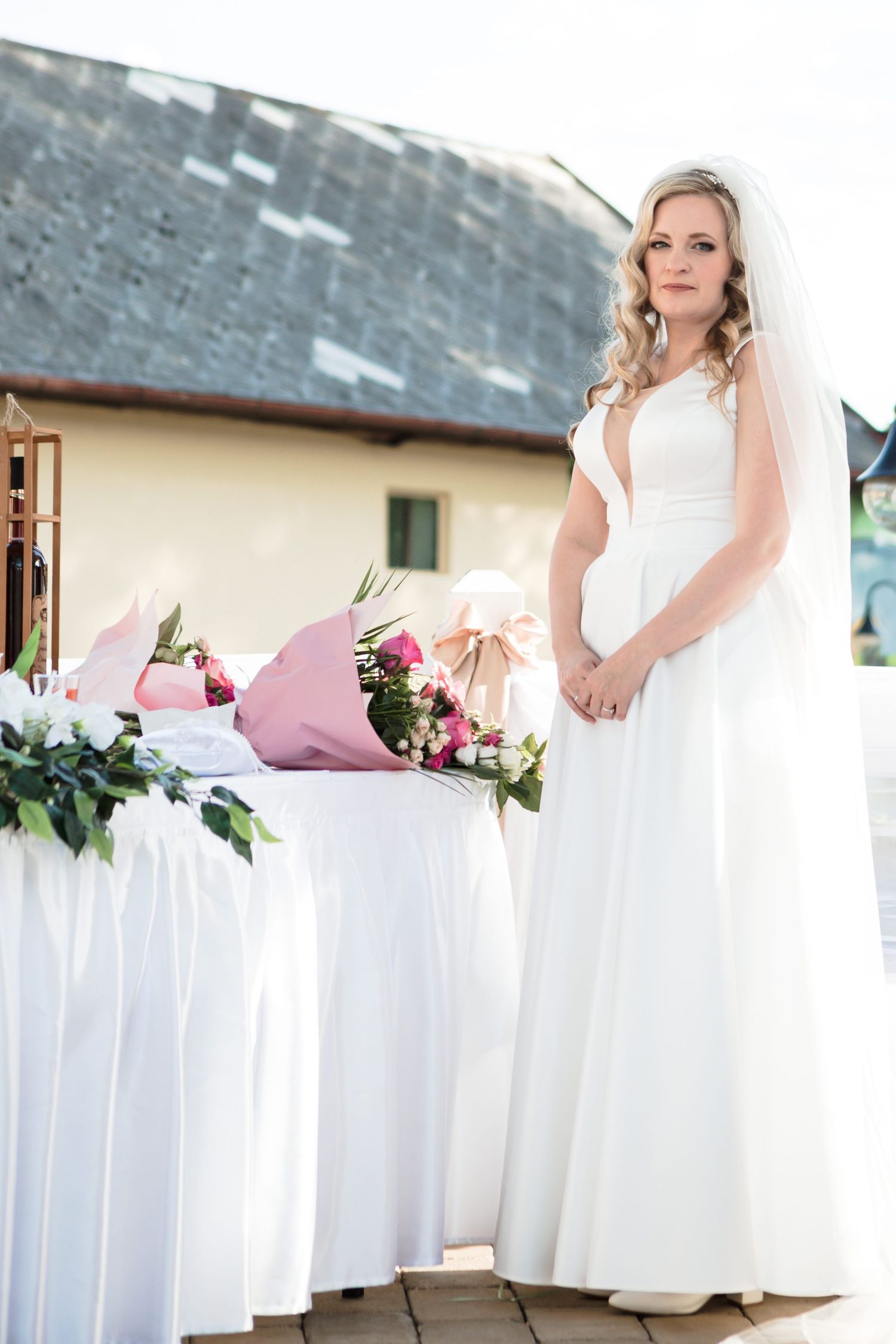 A bride in an elegant white wedding gown and veil stands beside a decorated table with pink floral arrangements.