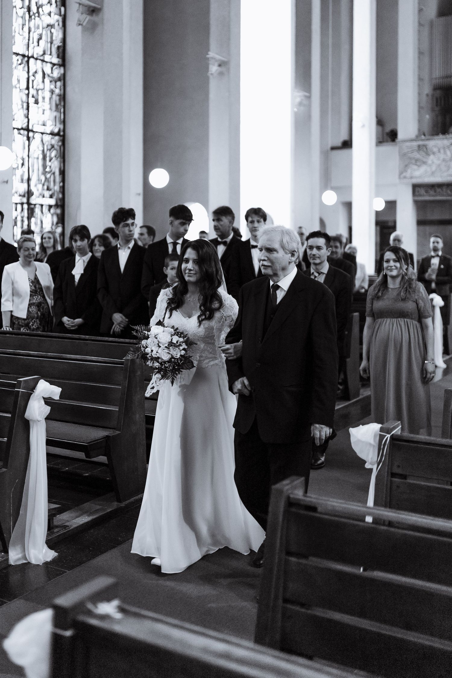 Wedding party members stand in church pews during a traditional ceremony with white floral decorations.