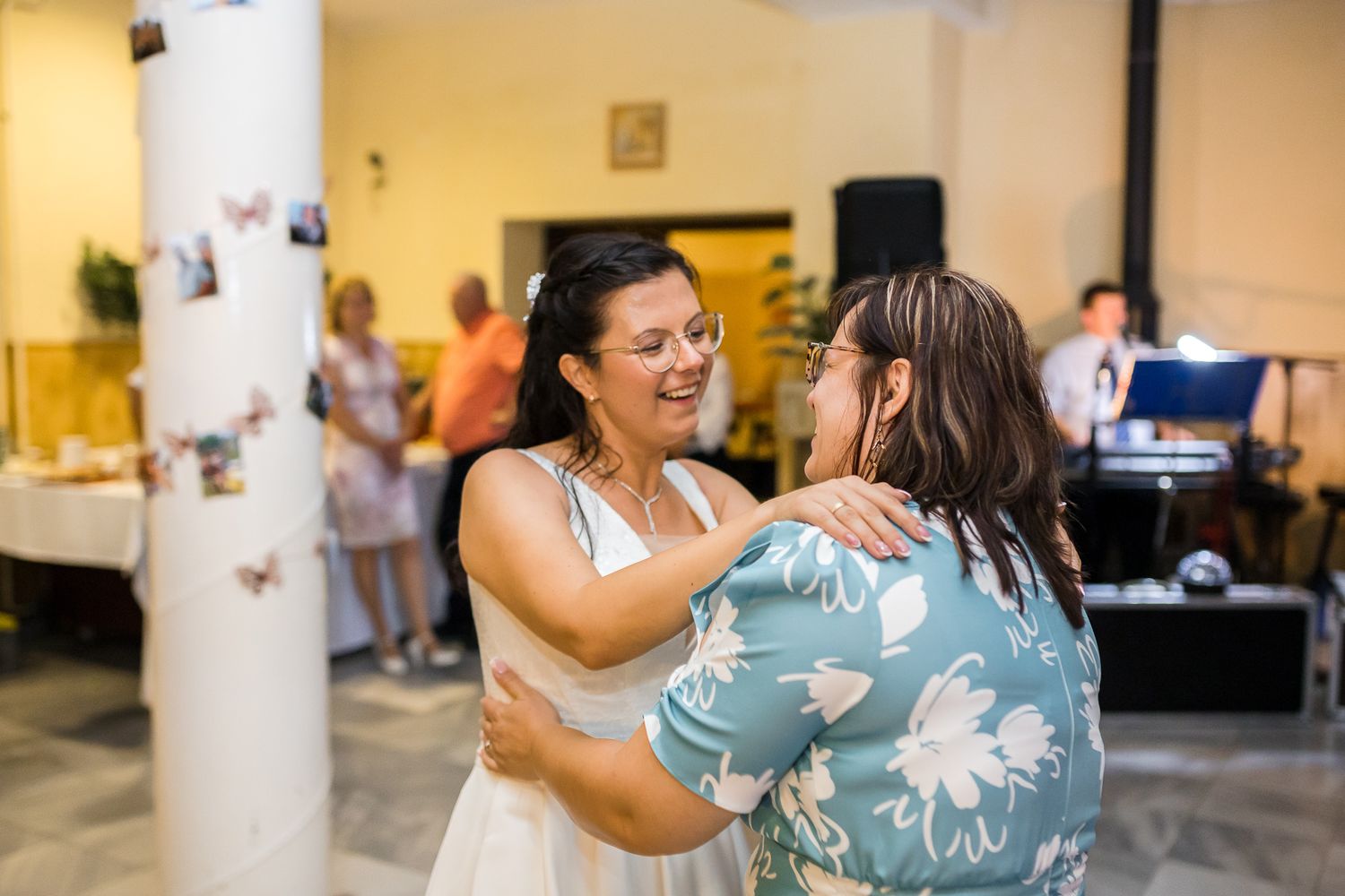 Two people share a joyful embrace while dancing at an indoor event with others visible in the background.
