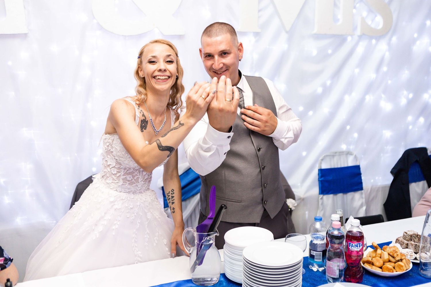 A couple in wedding attire sharing cake at their reception with a white backdrop and blue table decor.