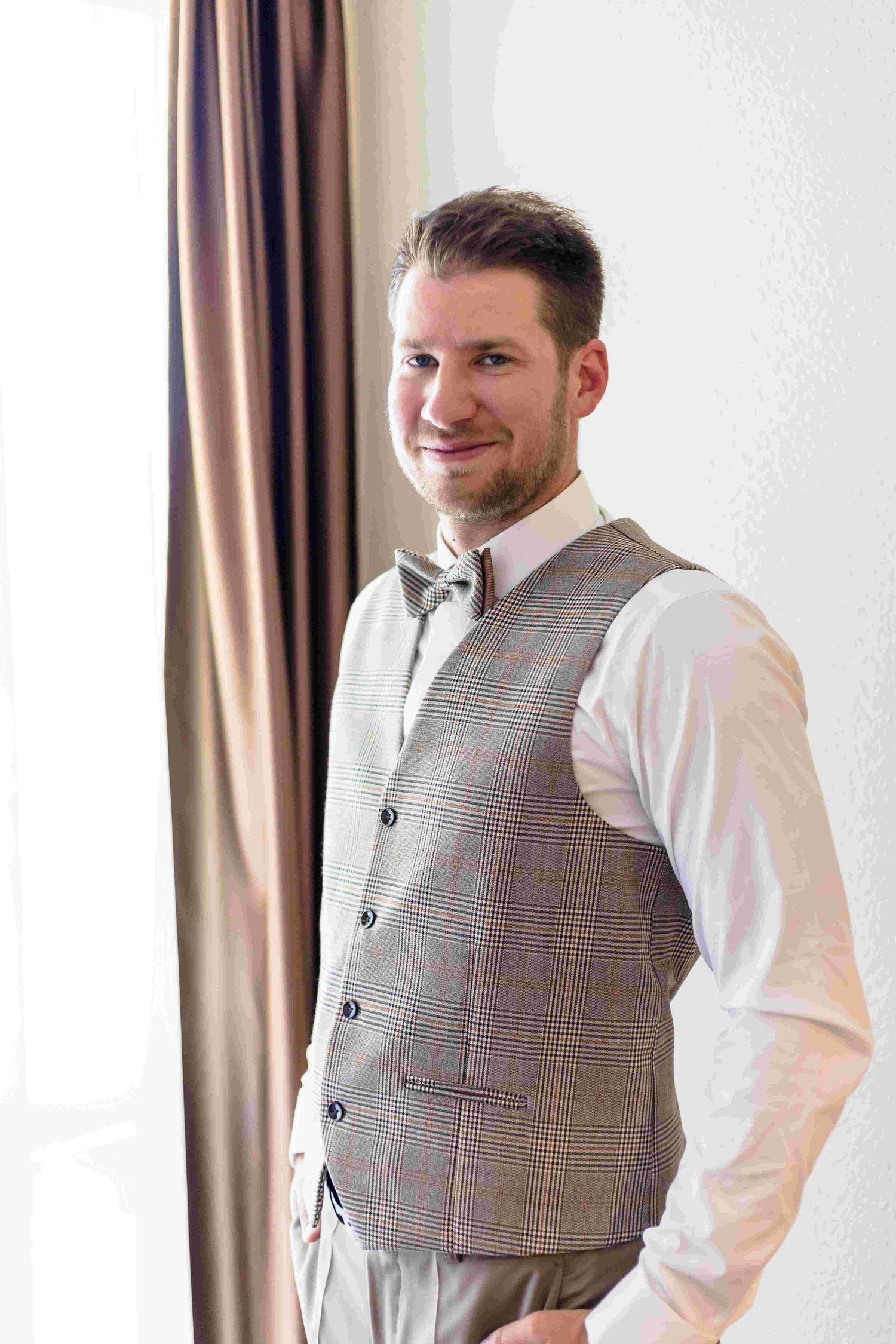 A person wearing a plaid gray vest and white dress shirt smiles while standing near curtains.