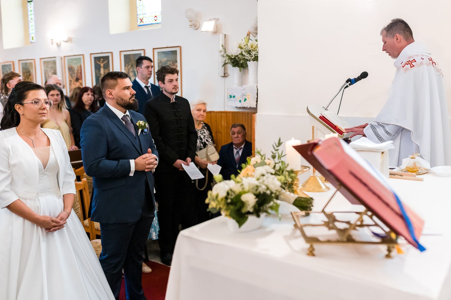 A wedding ceremony in progress with guests seated in a bright indoor venue decorated with white floral arrangements.