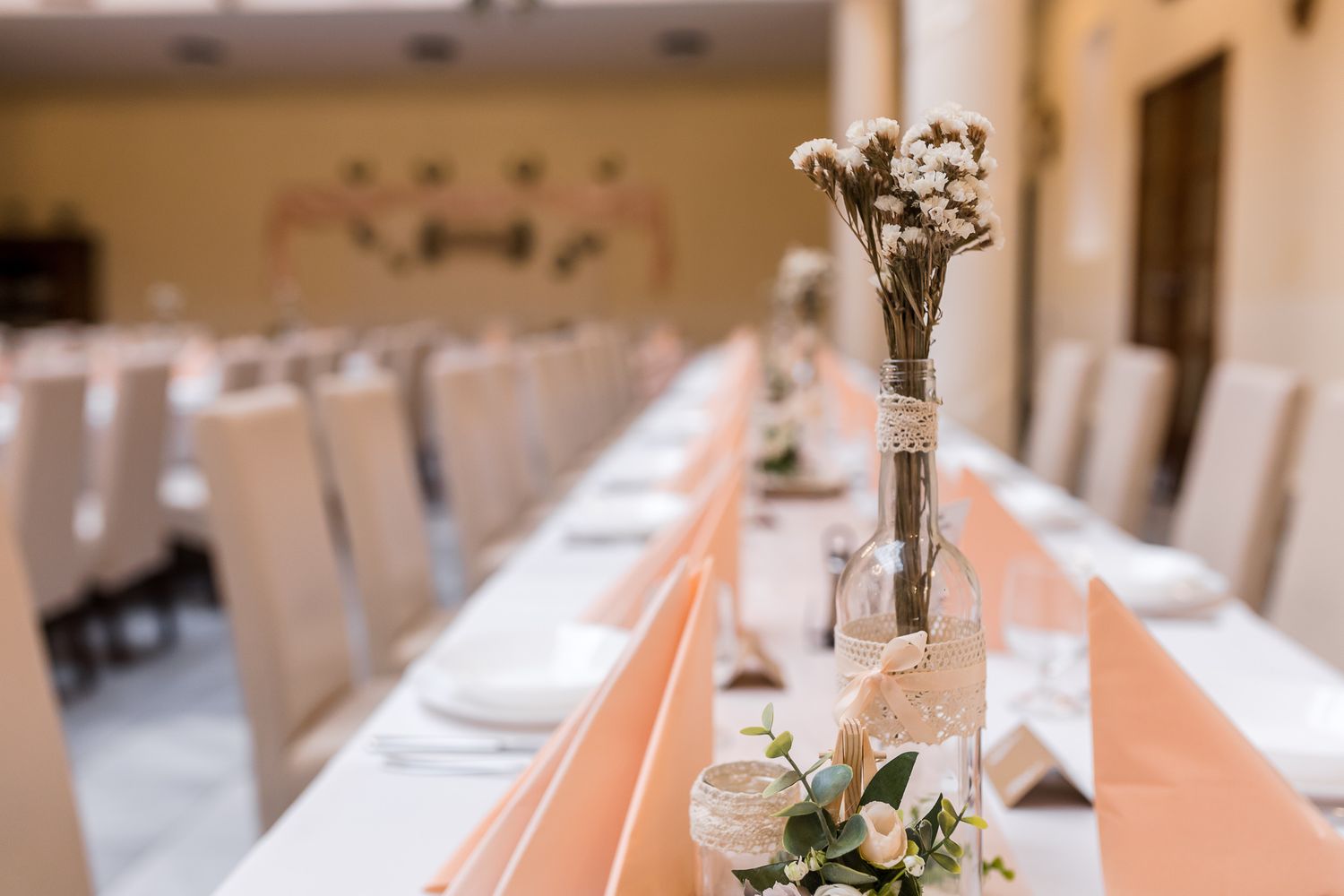 Elegant wedding reception tables decorated with white linens, peach accents, and floral centerpieces in a bright venue.