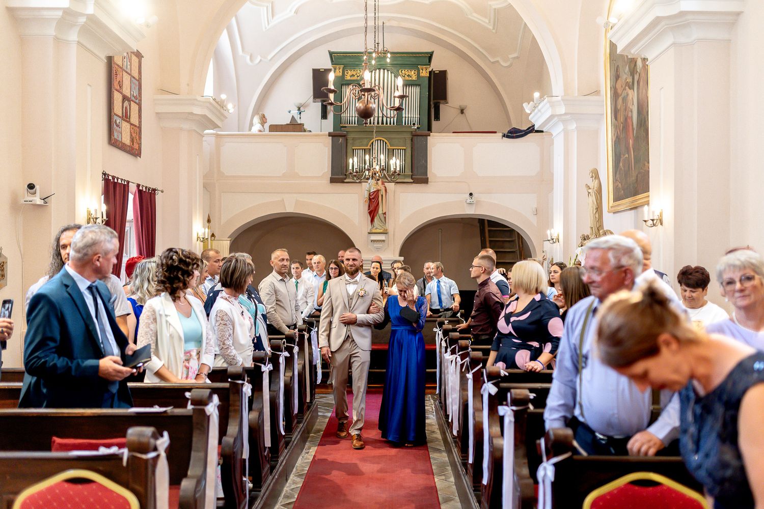 Wedding guests seated in white church pews during a ceremony beneath ornate arched ceilings and chandeliers.