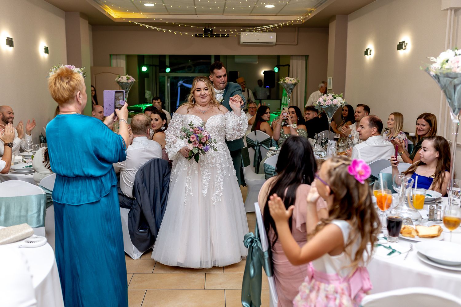 Wedding reception guests dancing and celebrating in an elegant venue with soft lighting and floral decorations.