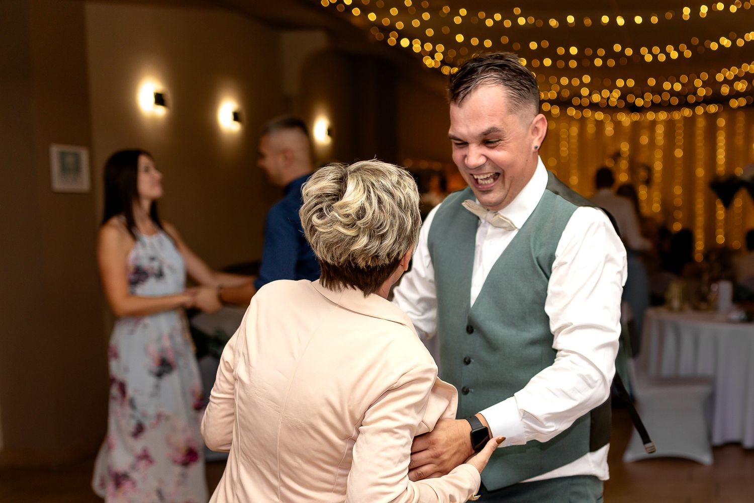 Wedding guests dance under twinkling string lights at an indoor reception venue.