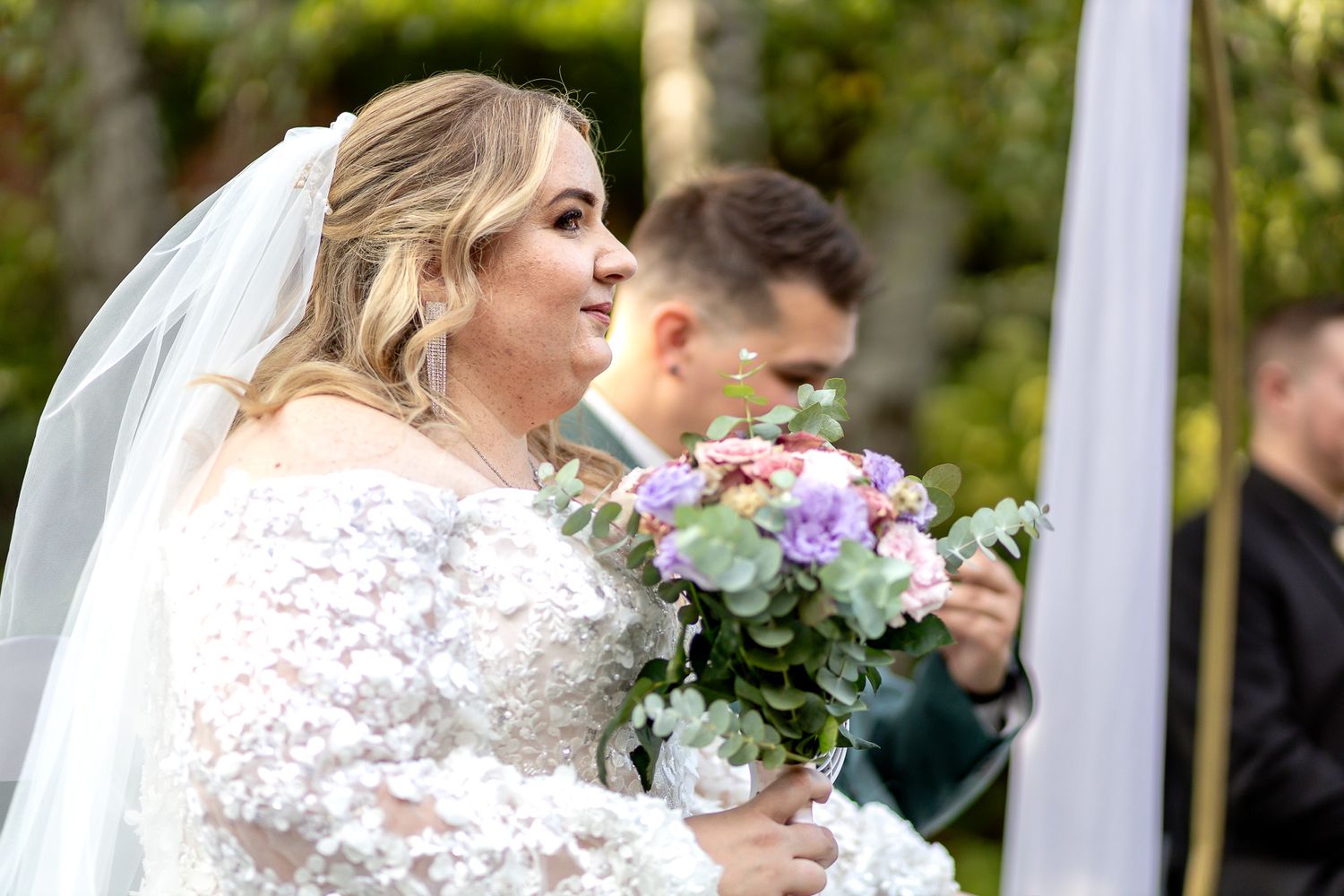 A bride and groom share a tender moment during their wedding ceremony under a white canopy outdoors.