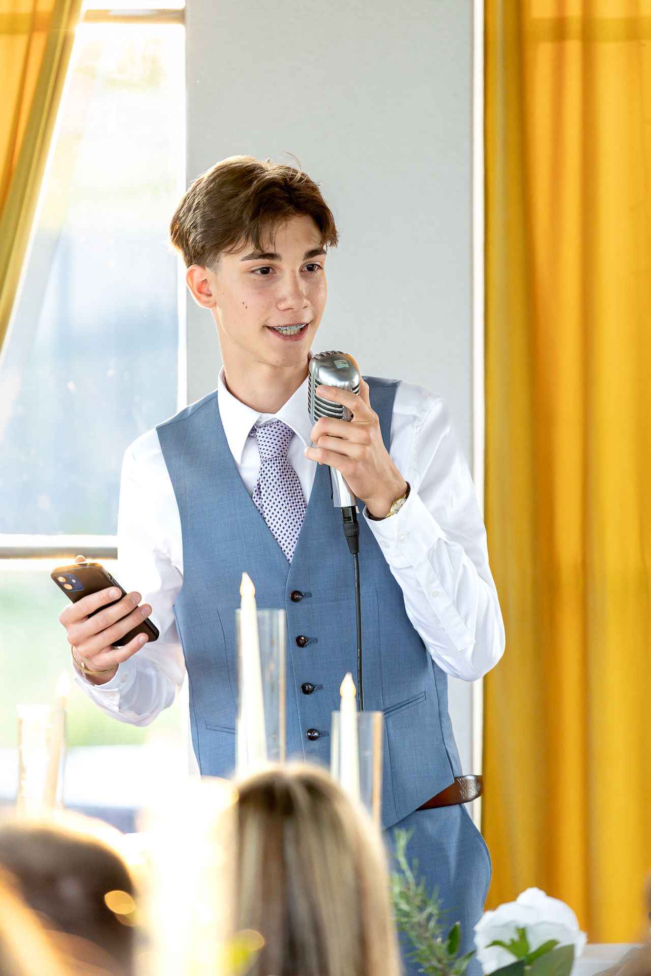 Person in formal attire giving an emotional speech at a wedding reception with yellow curtains.