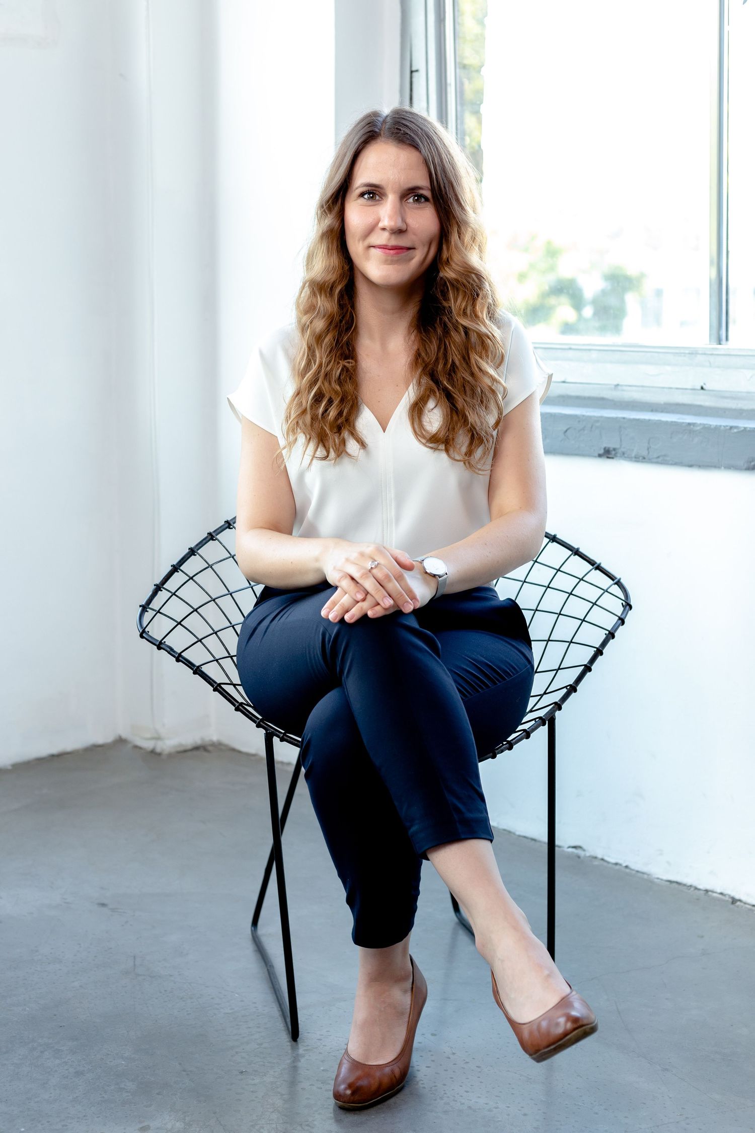 Person seated in modern wire chair wearing white top and navy pants in bright studio setting.