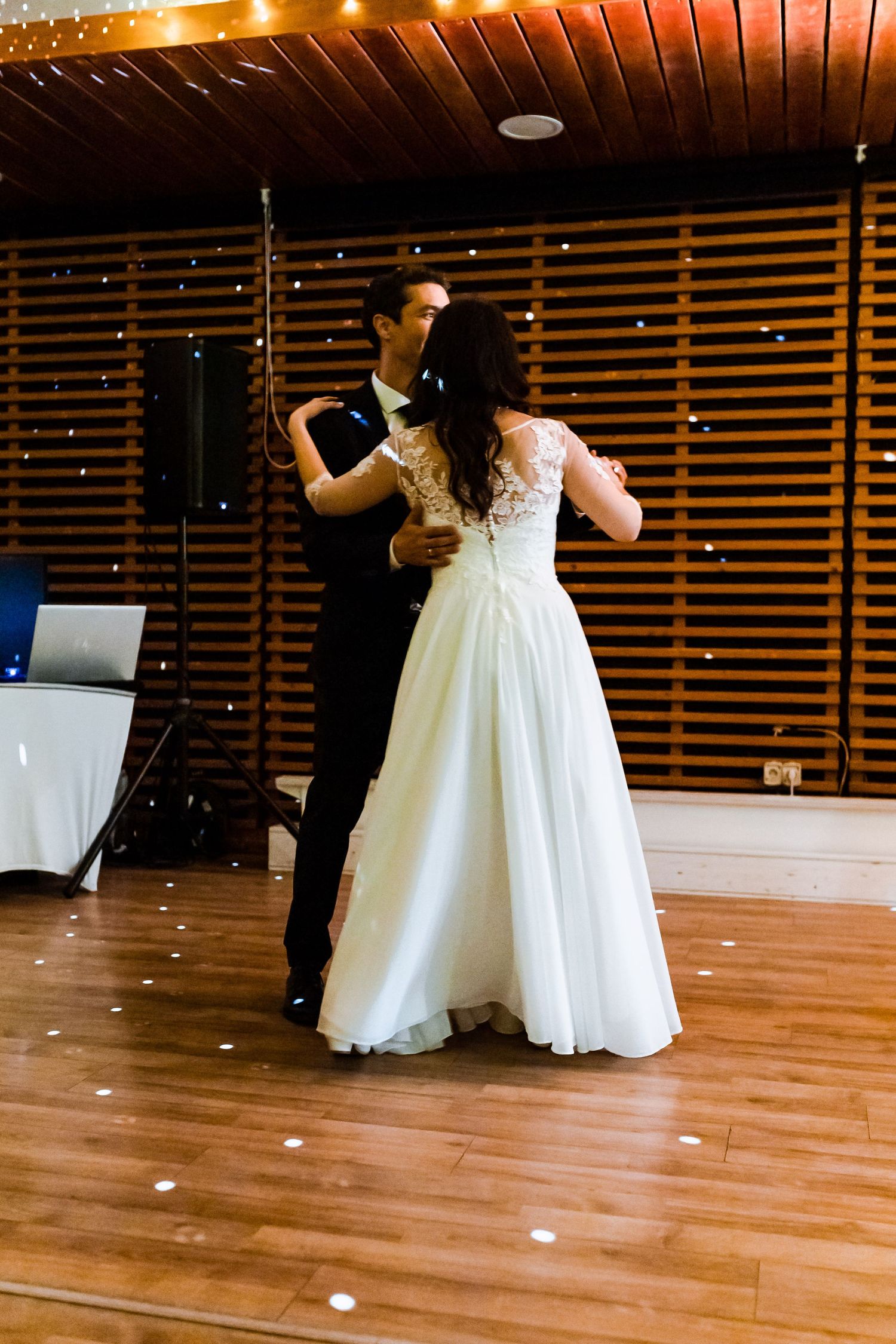 A couple in formal attire dances together on a wooden floor during what appears to be a wedding reception.