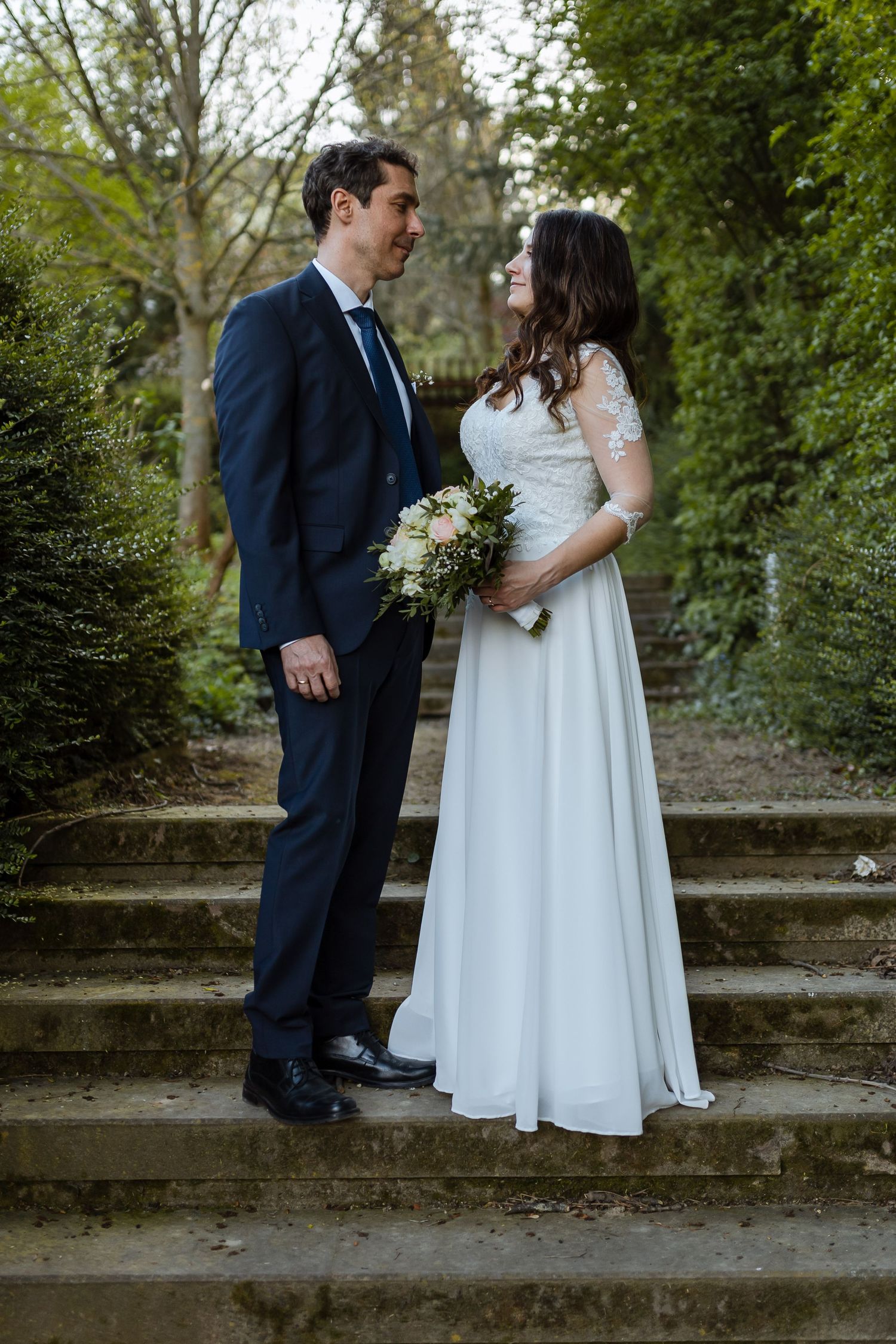 A couple in wedding attire shares romantic moments on stone steps surrounded by greenery in a garden setting.