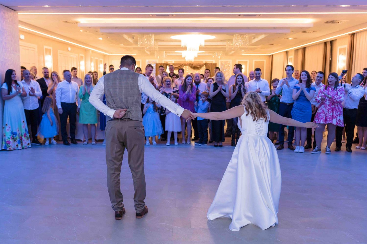 Wedding couple performs their first dance while guests in blue attire watch from the perimeter of a reception hall.
