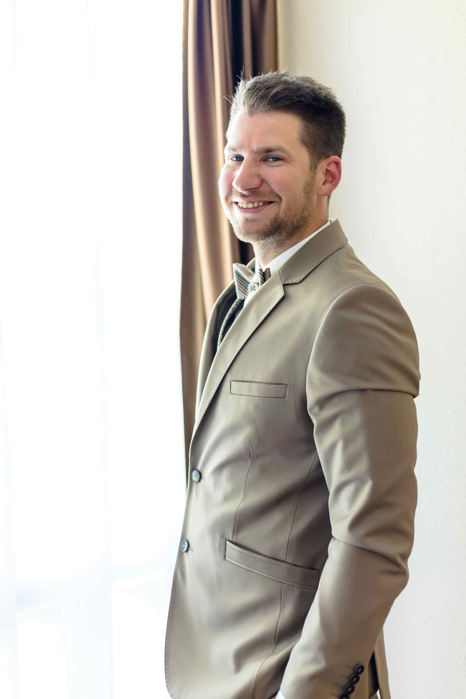 A person wearing a light beige suit jacket stands by a window in three different poses.
