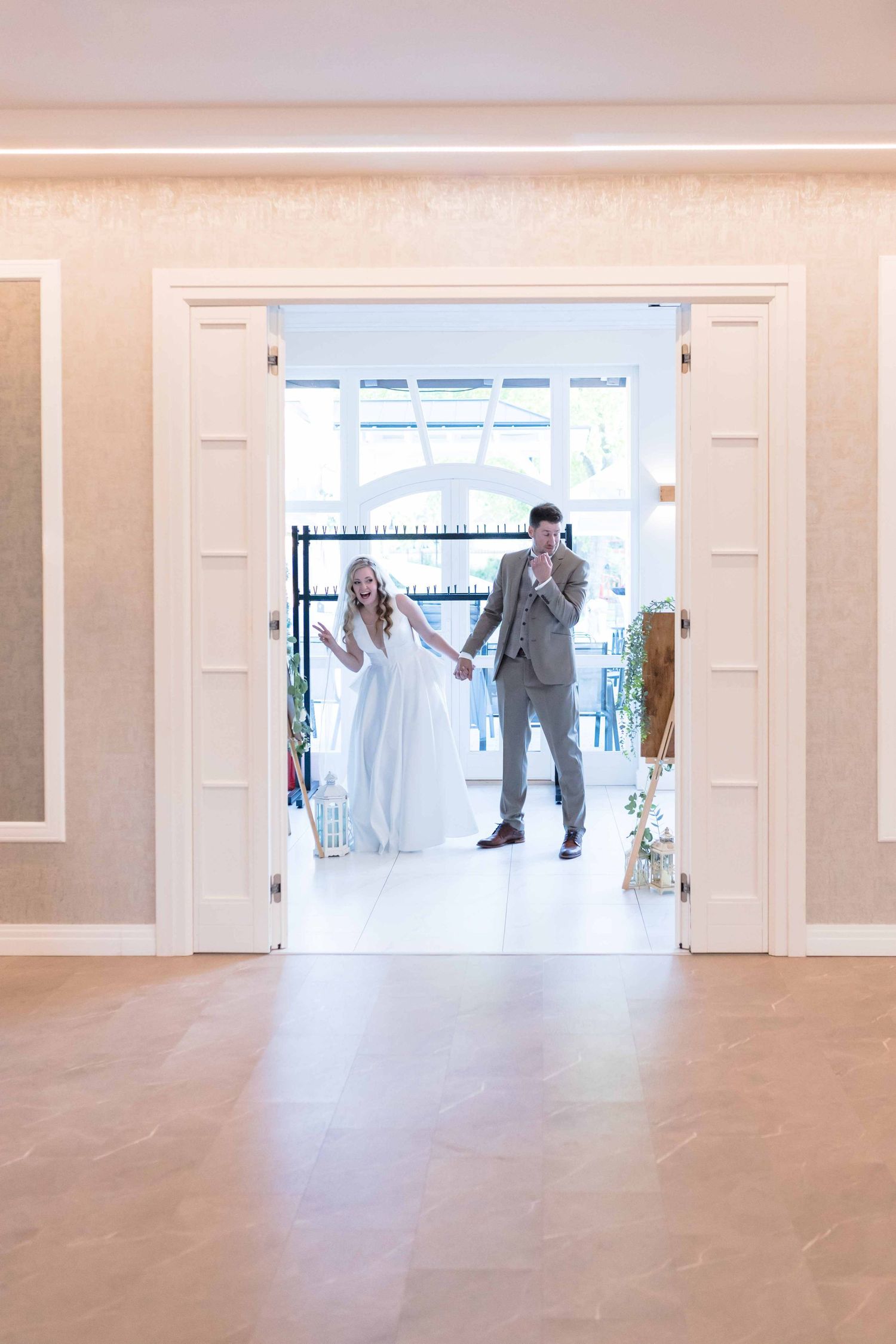 A couple in wedding attire stands in a bright doorway of an elegant white room with polished floors.