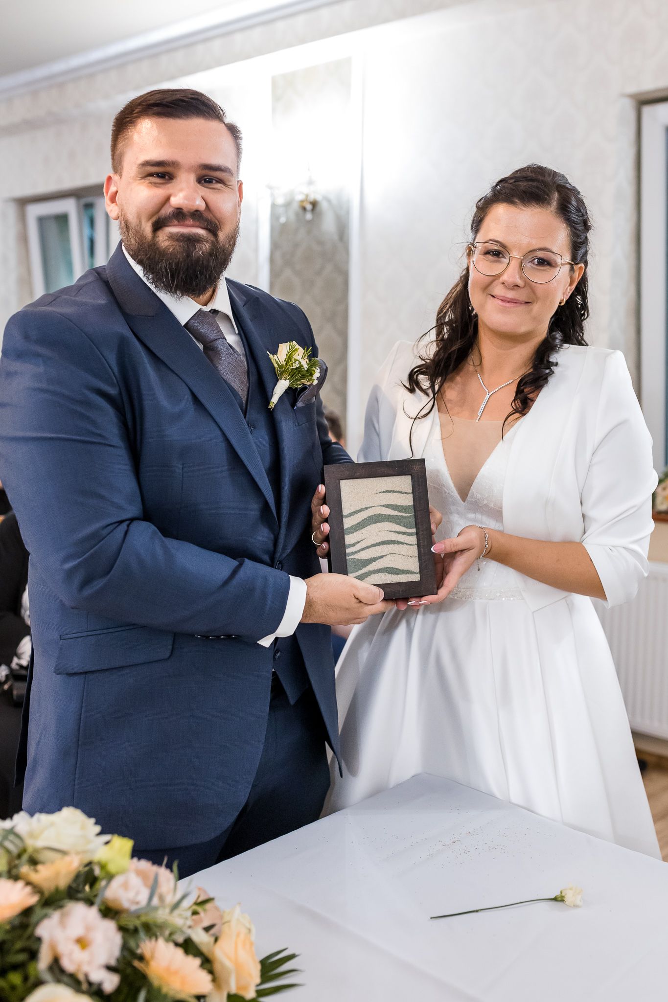 A couple at their wedding ceremony holding a decorative frame together, with floral arrangements on the table.