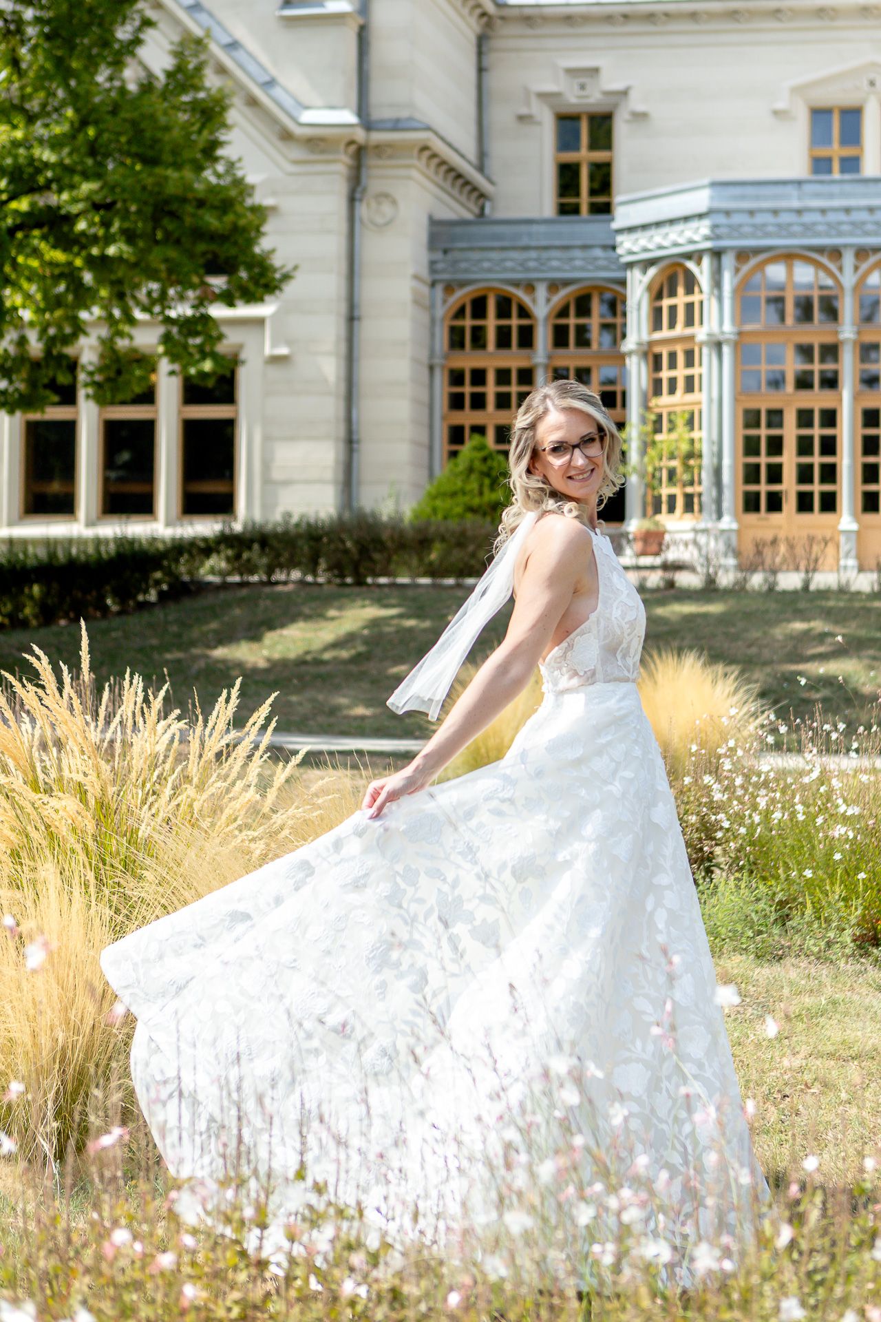 A flowing white wedding gown twirls gracefully in a sunlit meadow against the backdrop of a grand historic mansion.