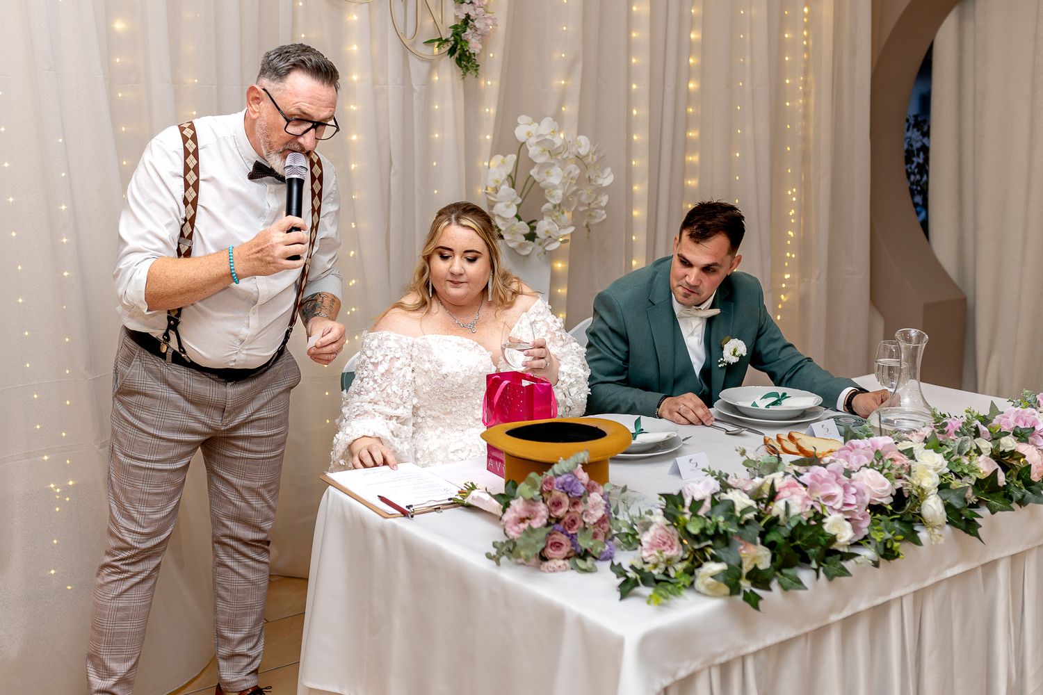 Wedding guests celebrate at a reception with speeches and toasts while seated at decorated tables with floral centerpieces.