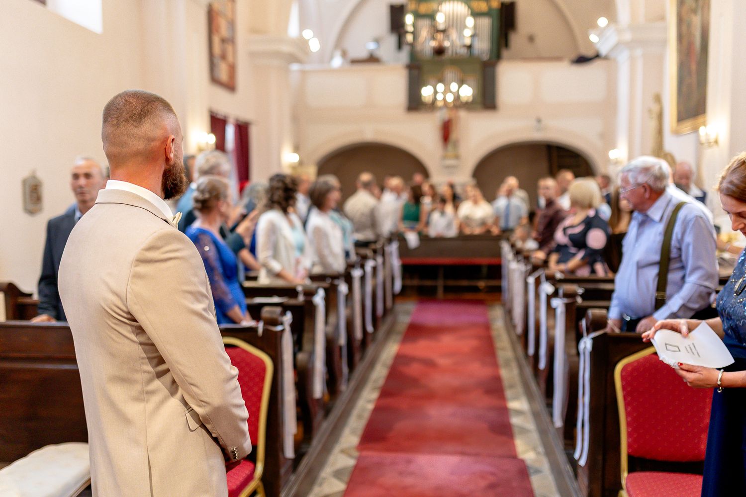 Wedding guests seated in church pews with red carpet aisle runner during indoor ceremony.