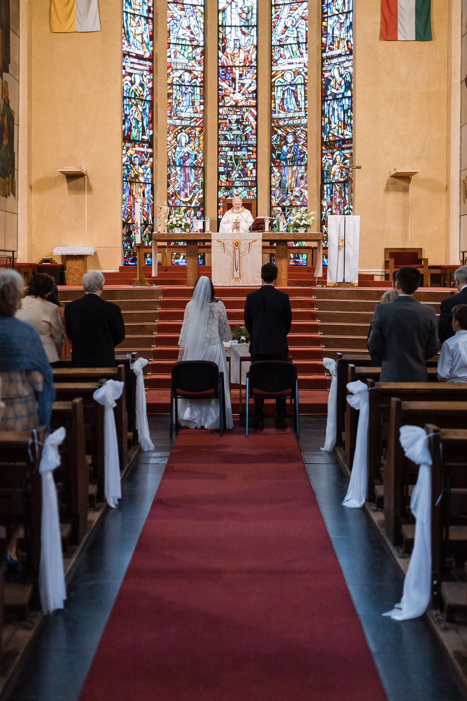 Inside a church with stained glass windows and red carpeting, white bows decorate the wooden pews for a wedding ceremony.