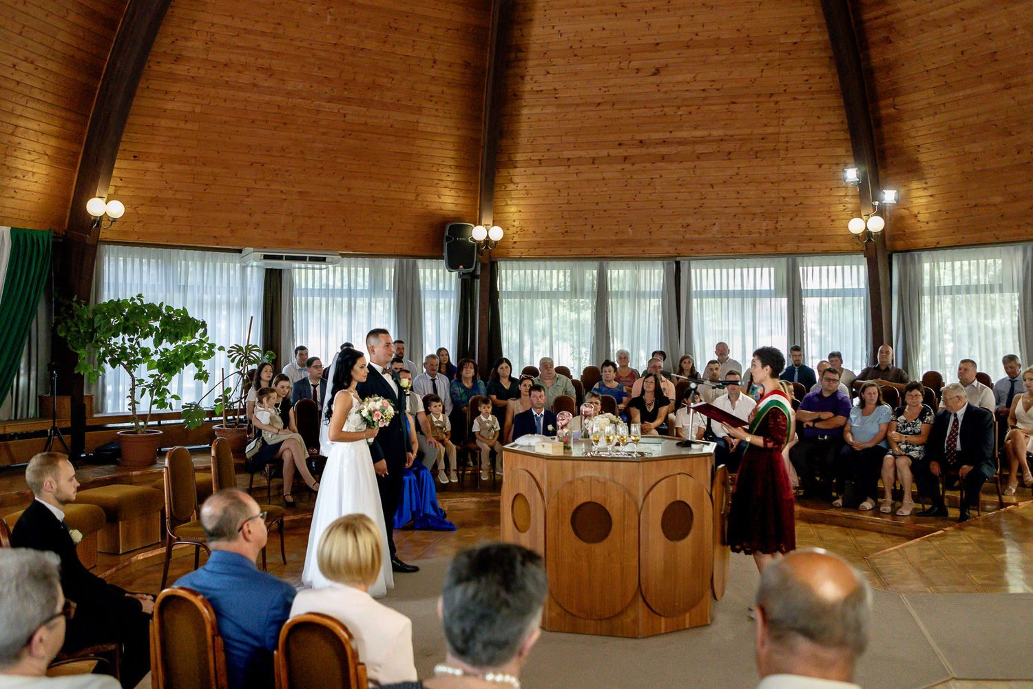 Panoramic view of a wedding ceremony in a modern wooden church with large windows and natural lighting.