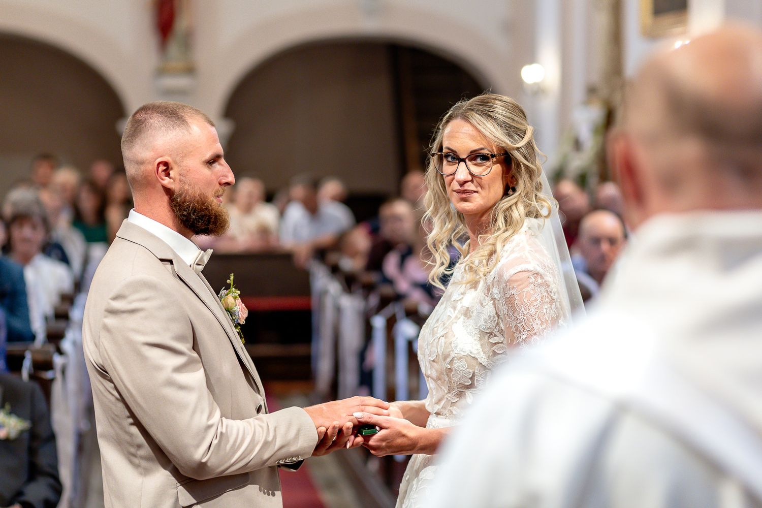 A bride and groom exchange rings during their wedding ceremony while guests look on in the background.