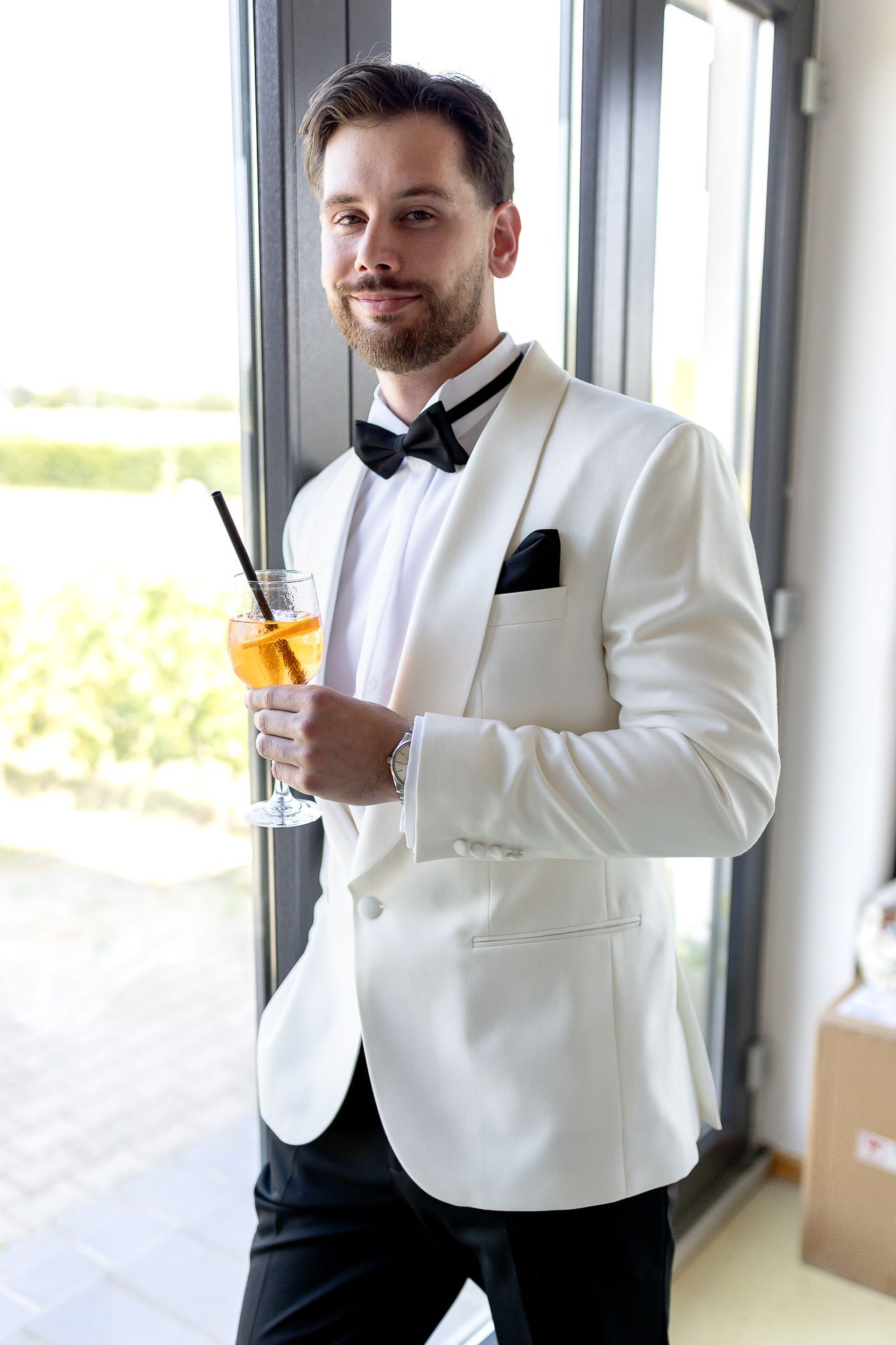 Two people in white tuxedos and black bow ties share drinks by a window.