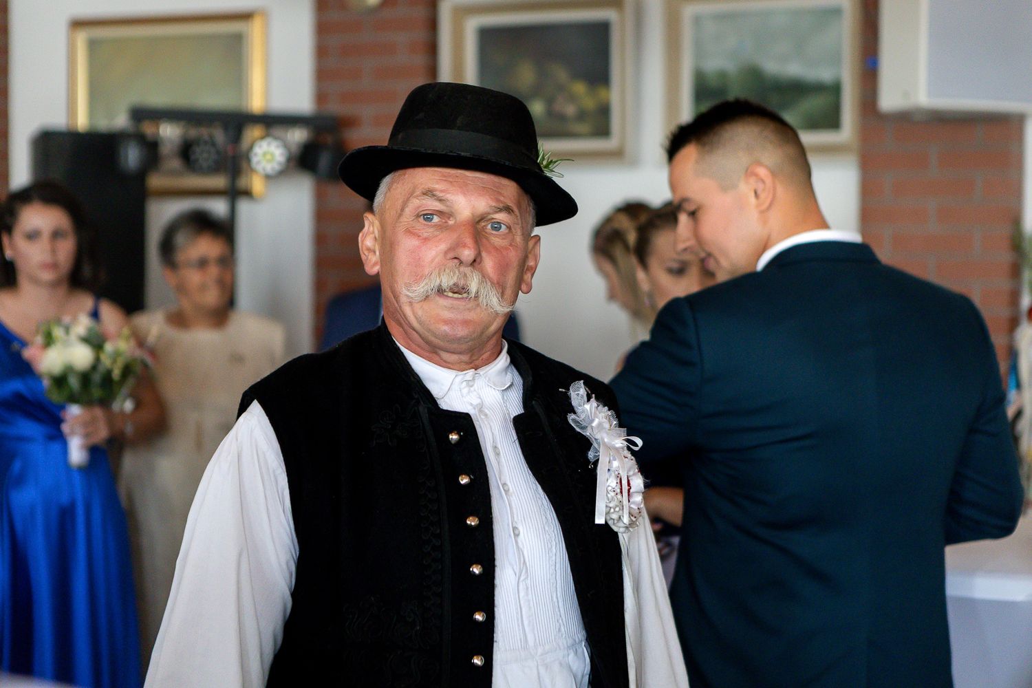 Traditional European folk costume with black vest and hat at a formal celebration with people in background.