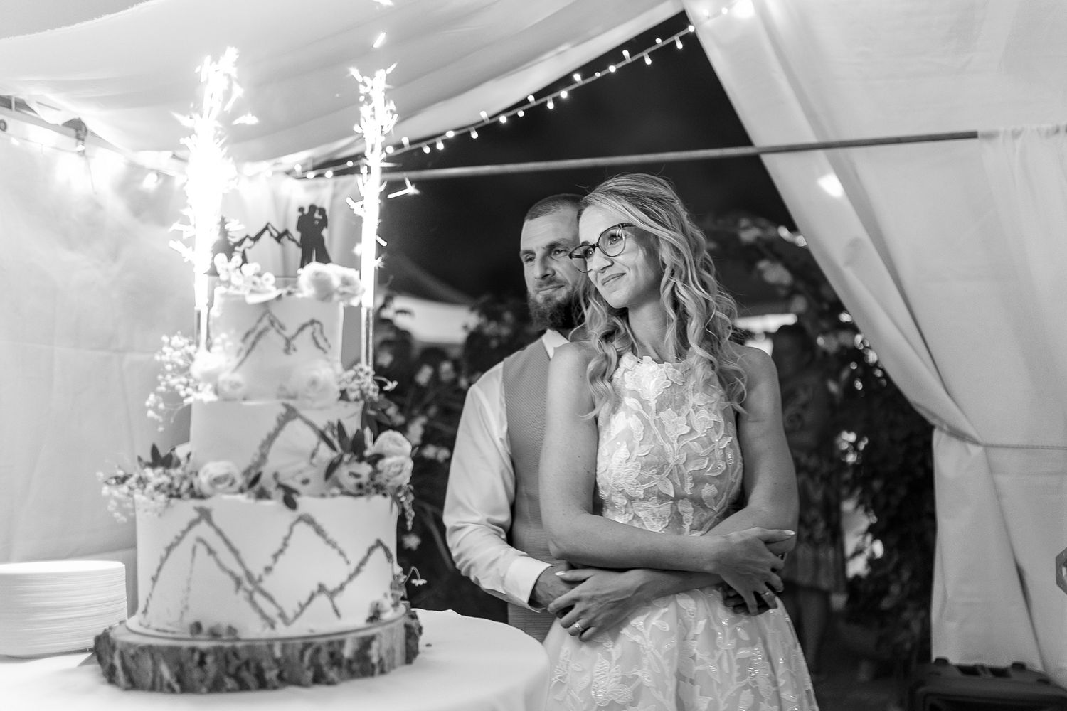 A romantic wedding couple shares a tender moment under a white tent decorated with twinkling lights.