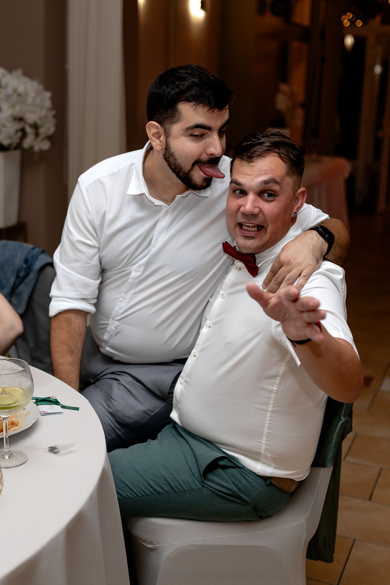 Two friends in white shirts laughing and enjoying a casual conversation while relaxing in chairs at a gathering.