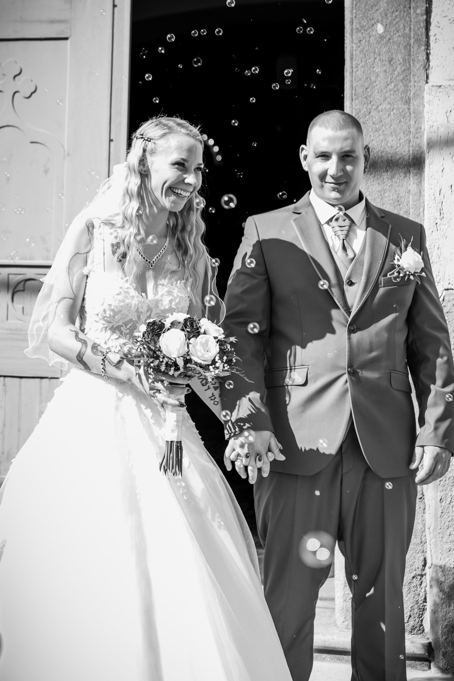 A wedding couple shares joyful laughter while being showered with bubbles outside a venue.