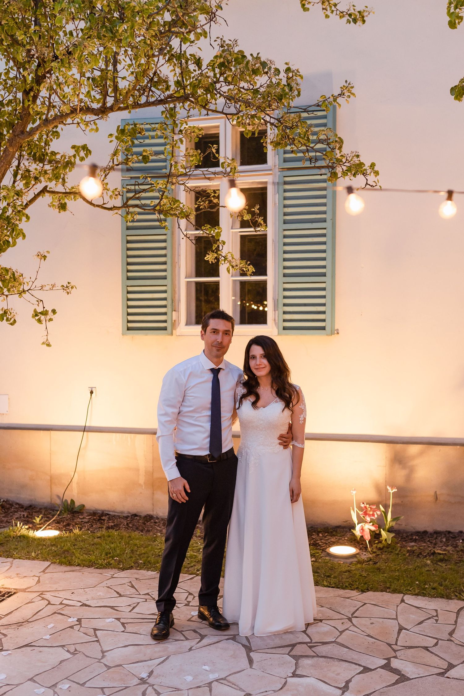 Couple poses together at sunset on stone patio with string lights and blue shuttered windows in background.